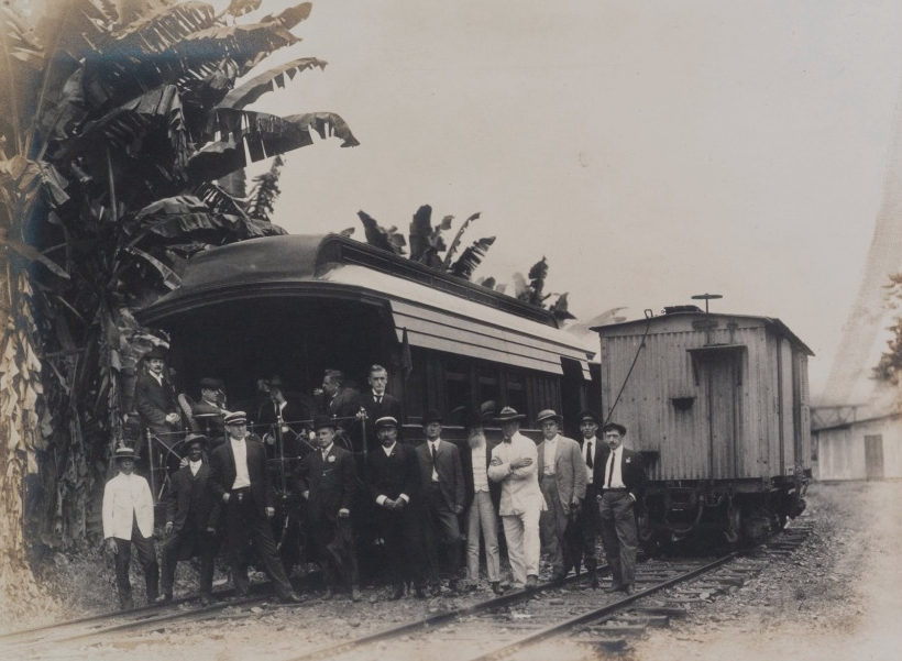 A group of men wearing suits and hats stand on and around an old train on railroad tracks. Tall plants and trees are visible in the background, and part of a building is seen to the right. The image is in sepia tones.