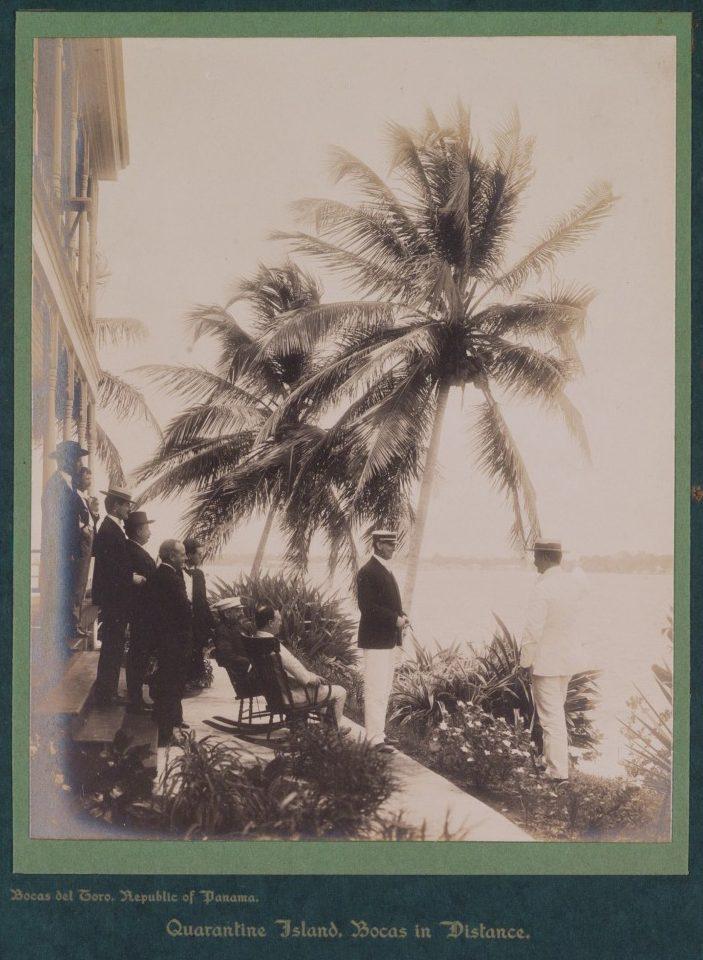 A vintage photograph shows a group of men in formal attire standing on a veranda adorned with lush tropical plants, overlooking the sea. Tall palm trees sway in the background, and a distant island is visible on the horizon.