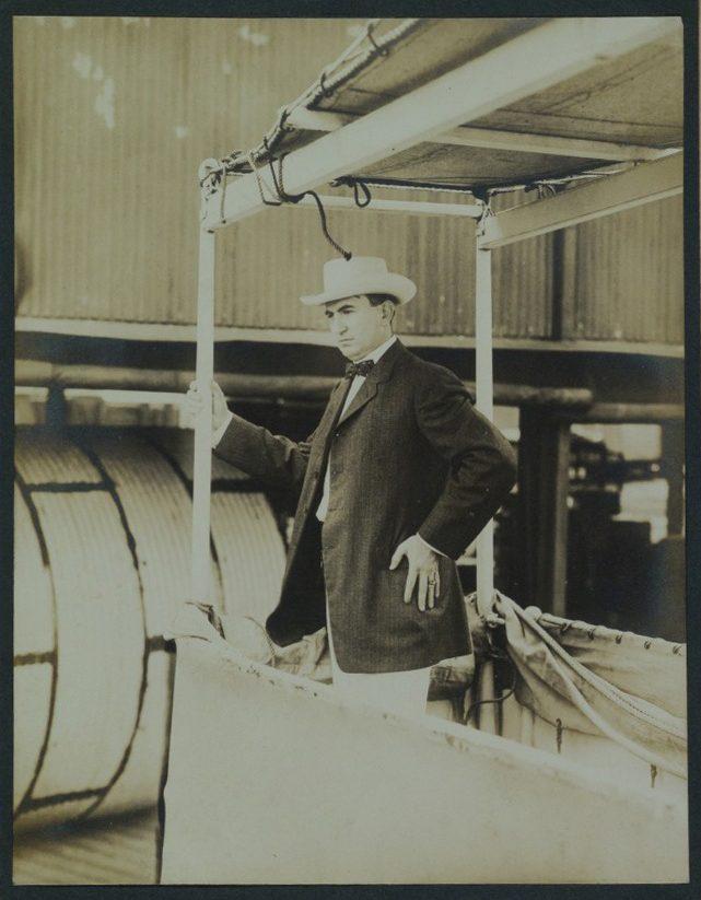 A man in a suit and white hat stands on a boat or dock, leaning against a rail. The background features industrial structures and large metal coils. The image has a sepia tone, giving it a vintage look.