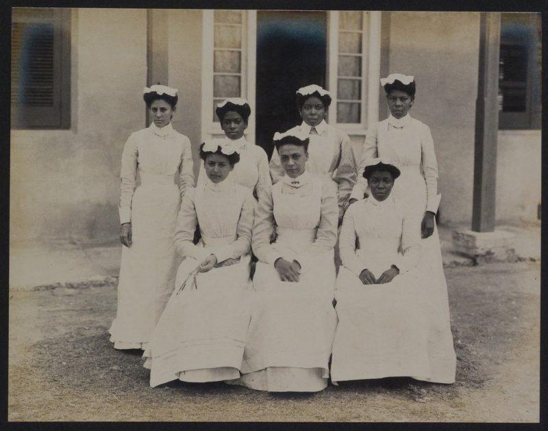 Seven nurses in white uniforms and caps pose for a group photo outside a building with windows and shutters. Four are standing in the back row, and three are seated in the front.