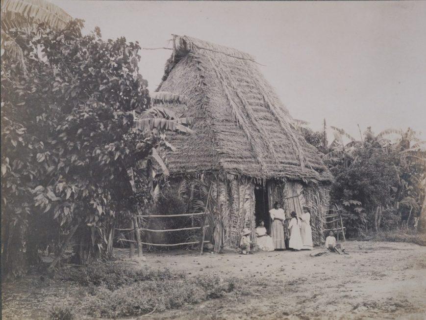 A vintage photograph of a group of people standing outside a thatched-roof hut surrounded by dense vegetation and banana trees. The scene appears rural, with a few individuals near the entrance and a child playing on the ground.