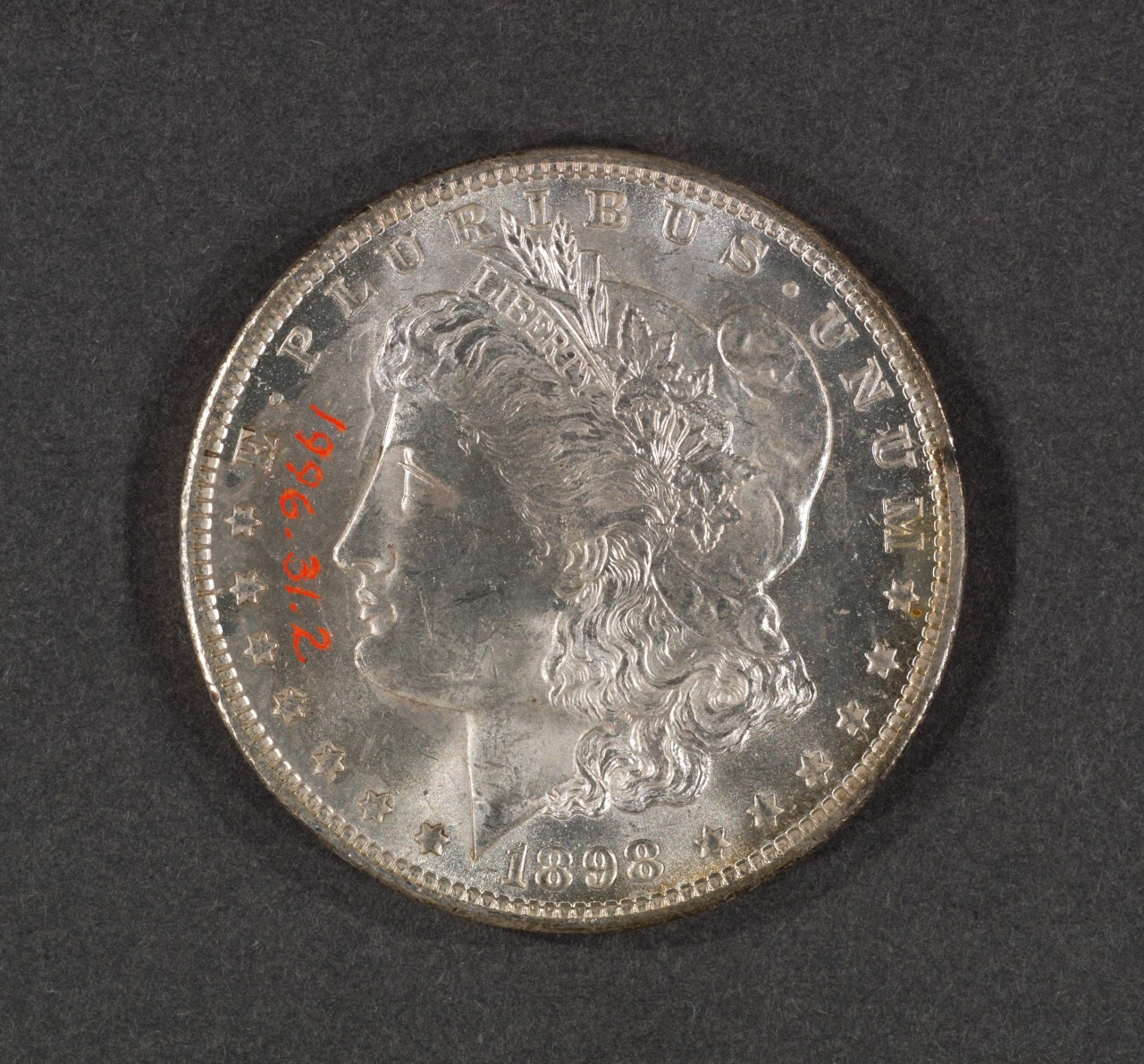 A silver coin with the profile of a woman wearing a headdress and the words E PLURIBUS UNUM along the top. The date 1898 is at the bottom. The number 1996.312 is in red on the side.