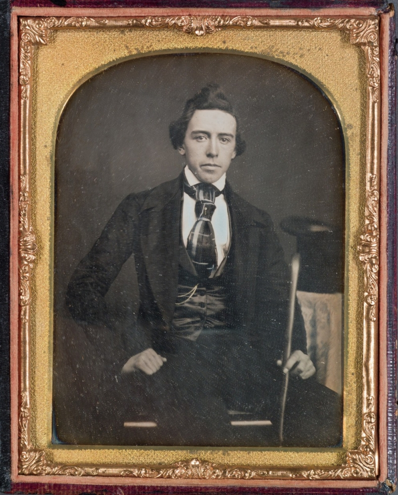A sepia-toned, vintage photograph of a man in formal 19th-century attire, seated and holding a cane. He wears a dark suit with a vest and cravat. The image is framed with an ornate golden border.