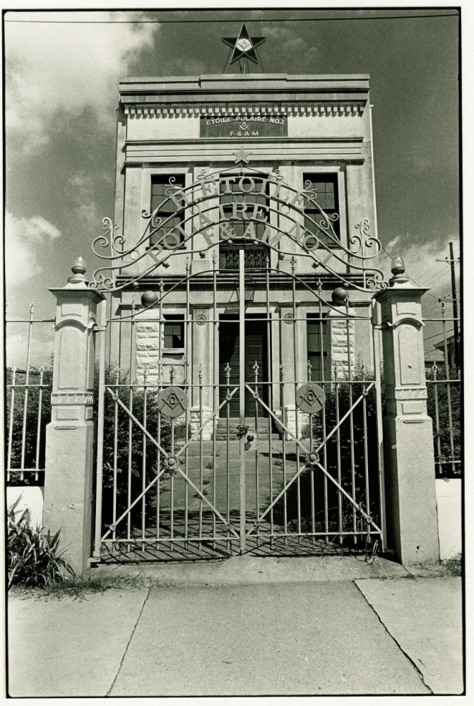 Black and white image of a historic stone building with a decorative wrought iron gate. The sign above reads Tolaga Bay & Districts RSA Memorial Clubrooms. A star is centered atop the structure. Cloudy sky in the background.