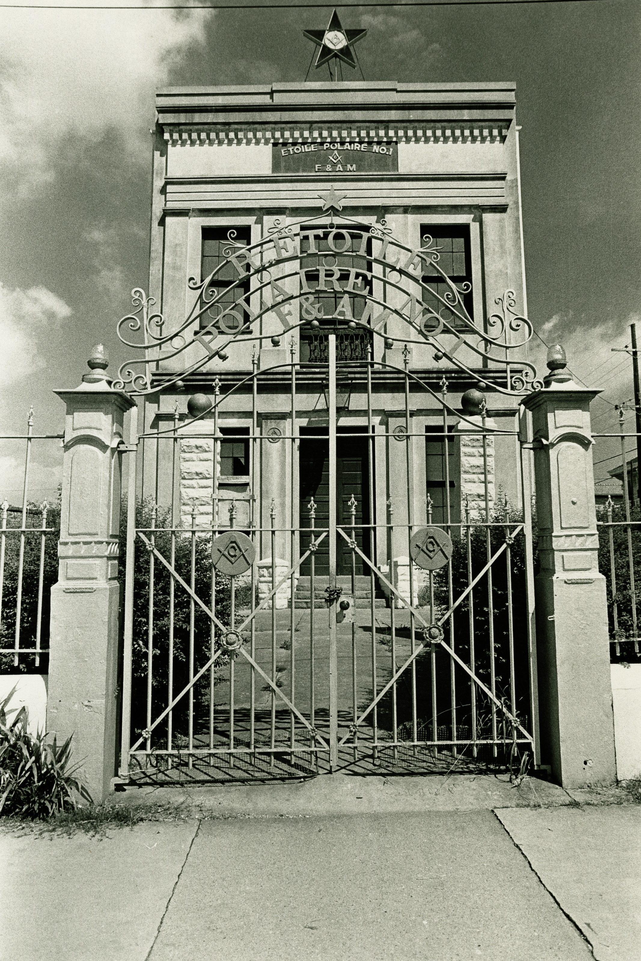 A black and white photo of an ornate iron gate in front of a historic stone building. The gate features decorative patterns and a central star design. The building has a sign above the entrance and is set against a partly cloudy sky.