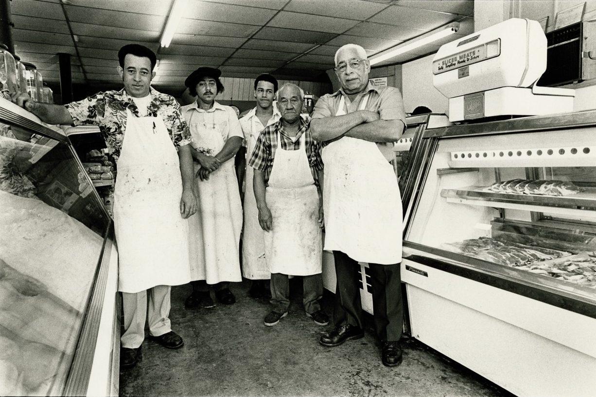 A black and white photo of five men wearing aprons standing in a butcher shop. They pose in front of display cases with meat products. The shop has tiled flooring and fluorescent lighting.
