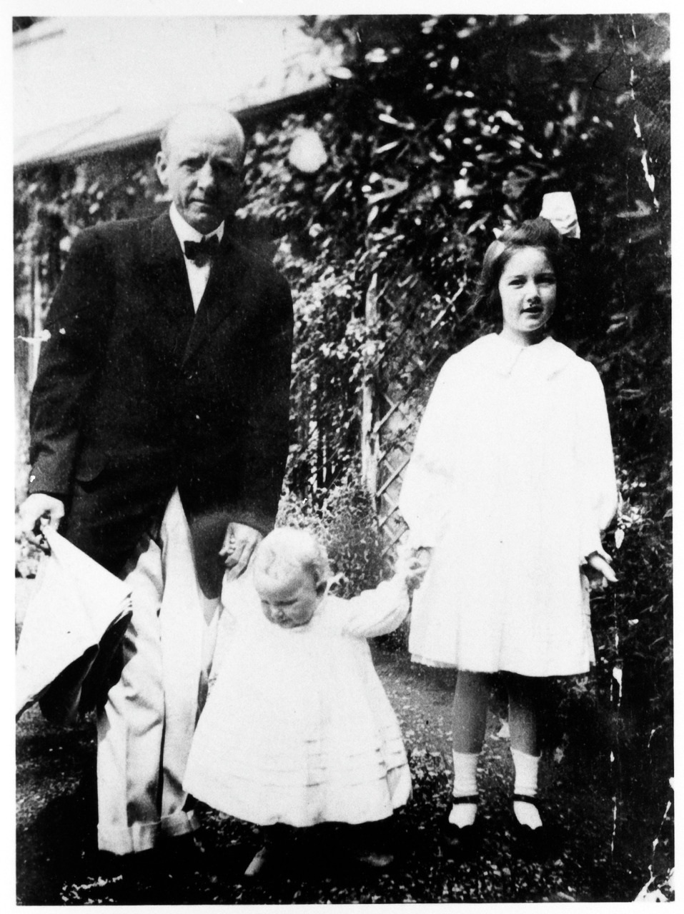 A black and white photo of an older man in a suit holding hands with two young children. The girl on the right wears a white dress with a large bow in her hair. The younger child in the center is also in a white outfit. They are standing outdoors.