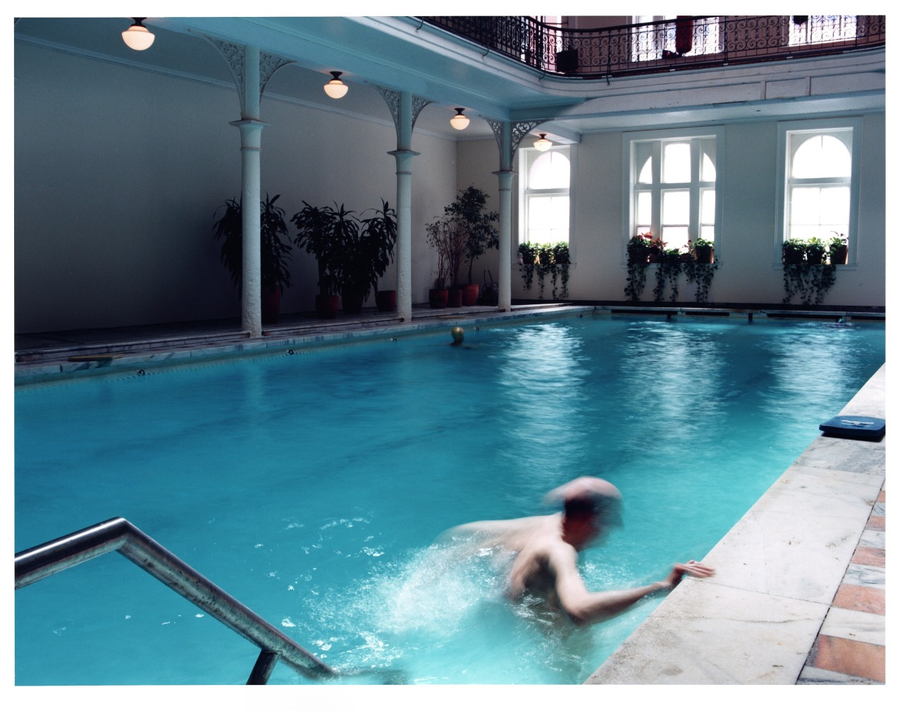 A person swimming in an indoor pool with vintage architecture. The pool is surrounded by white pillars, potted plants, and arched windows. The person is approaching the pools edge, creating a splash in the water.