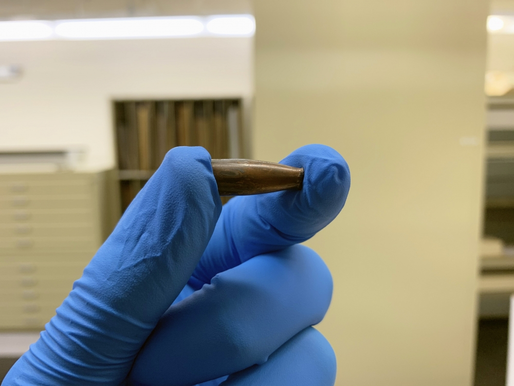 A person wearing blue gloves holds a single bullet between their thumb and index finger. The background is a blurred indoor setting with neutral colors.