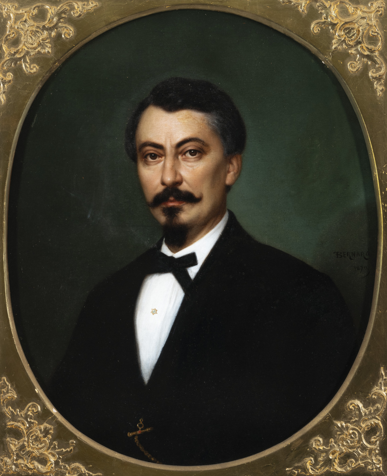 A man with dark hair and a mustache is depicted in a formal portrait. He wears a black suit, a white shirt, and a black bow tie. The background is dark green, and the ornate frame is gold.