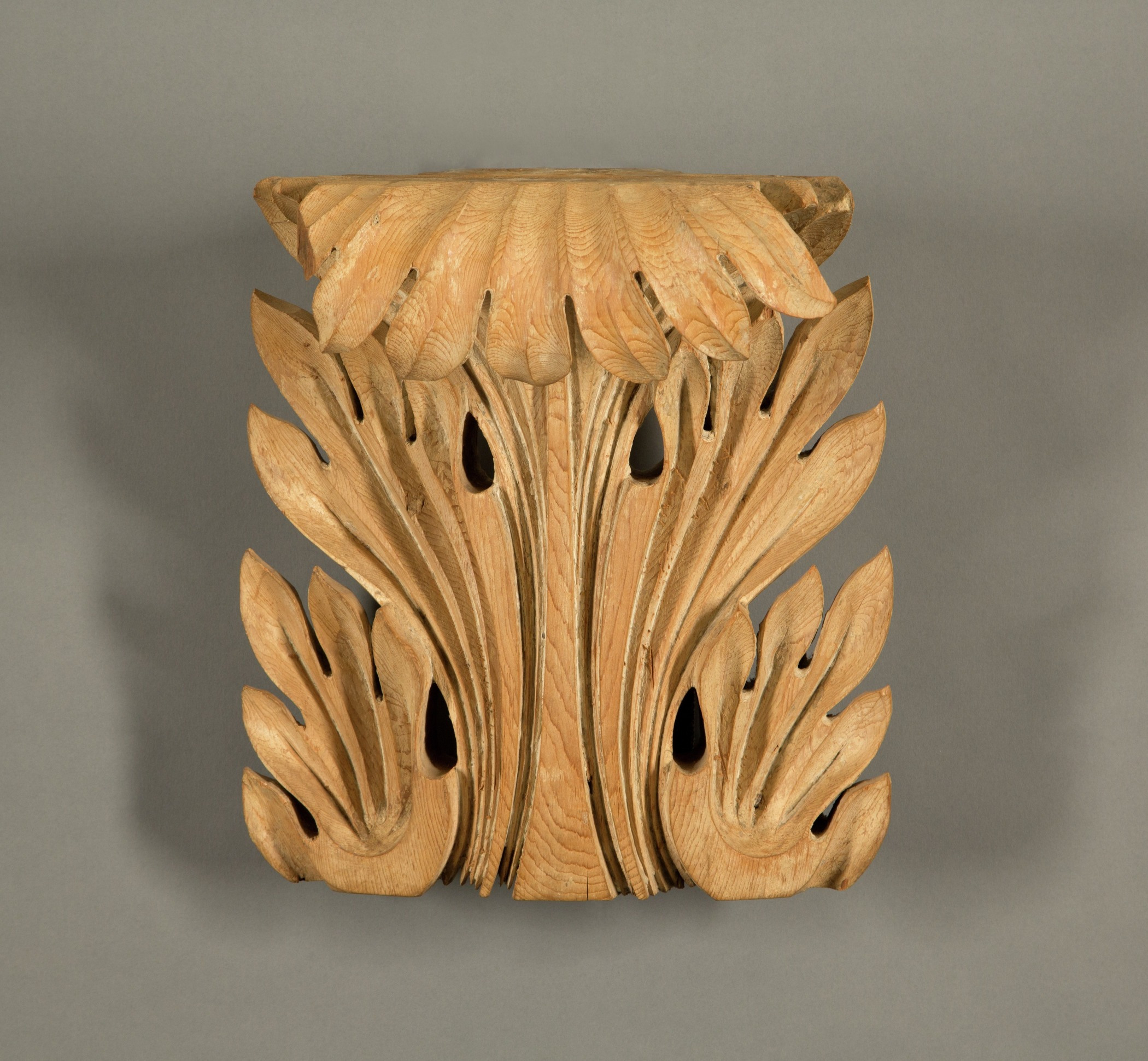 A decorative wooden wall bracket featuring intricately carved leaf and flower motifs. The design includes graceful curves and organic shapes, highlighting the natural wood grain. The piece is displayed against a plain gray background.