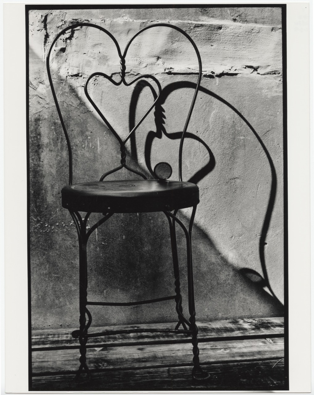 A black and white photo of a metal chair with a heart-shaped back. The chair casts a prominent shadow on a textured wall, creating a striking visual contrast. The shadow echoes the heart shape of the chairs backrest.