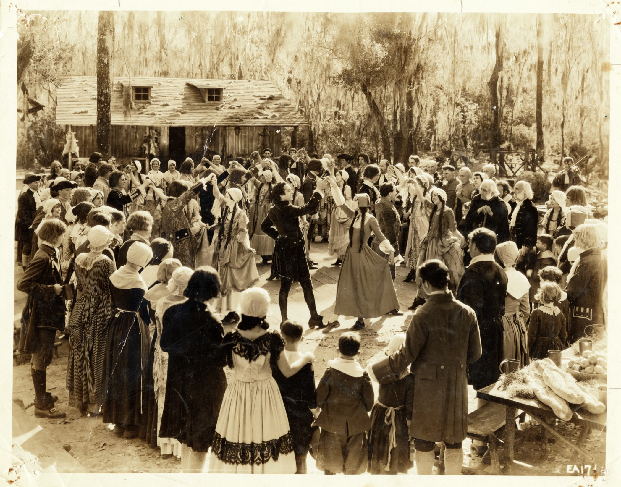A large group of people in historical costumes gather outdoors, forming a circle around dancers in the center. The background features trees and a rustic wooden building, creating a lively but vintage scene.