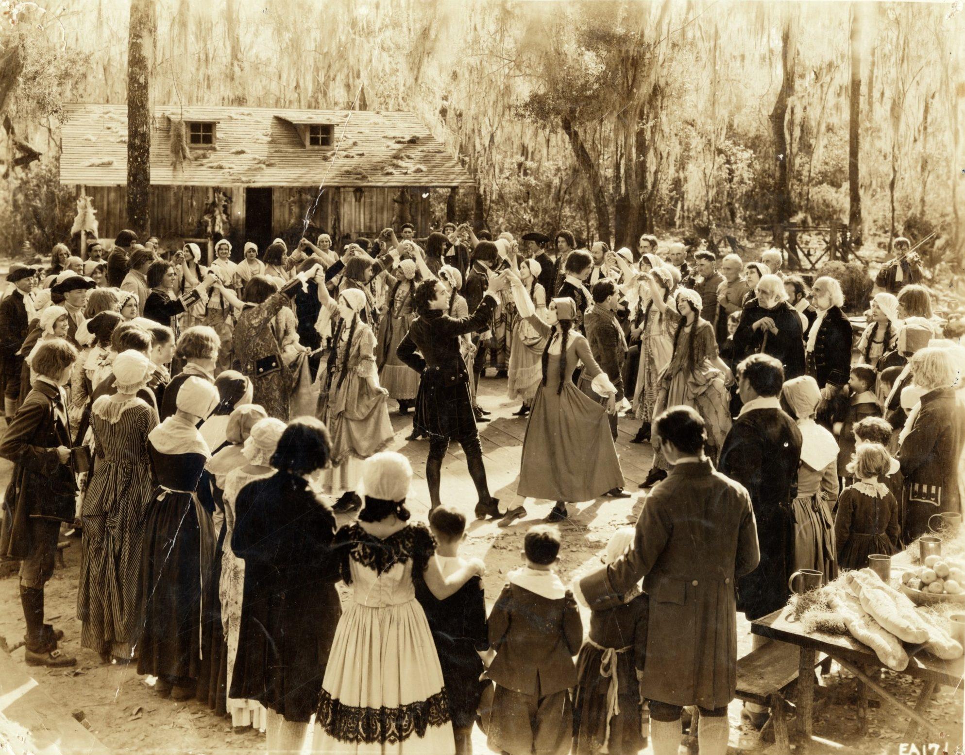 A vintage sepia-toned image shows a large group of people in historical costumes dancing in a circle outdoors. Spectators in period attire surround the dancers, with trees and a rustic wooden building in the background.