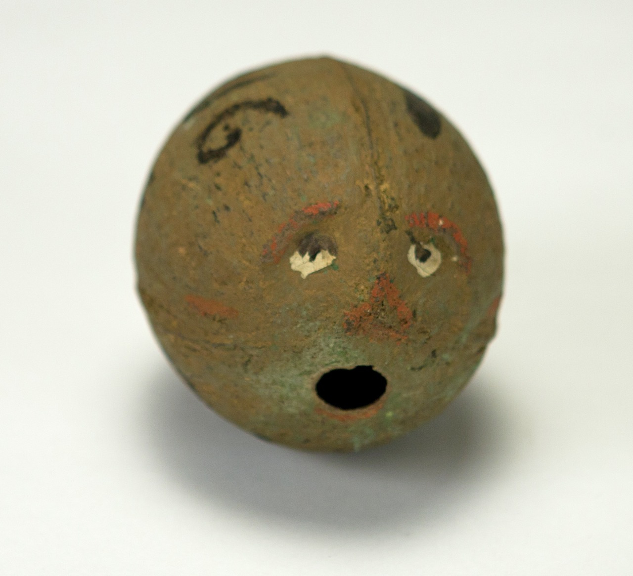 A round, brown object with a roughly painted face, featuring wide eyes and a small open mouth, is set against a plain background. Lines for eyebrows and cheeks are visible.