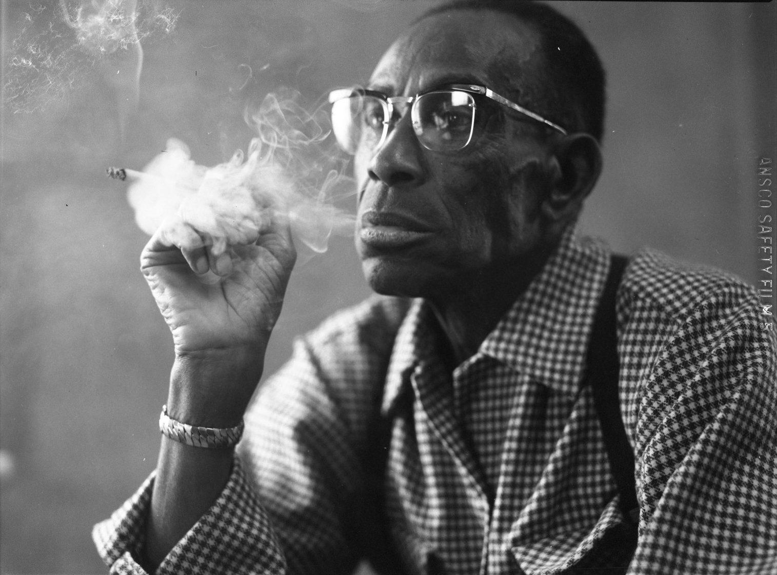 An older man wearing glasses and a checkered shirt is sitting and smoking a cigar. He looks thoughtful as smoke swirls around him in the black and white image.