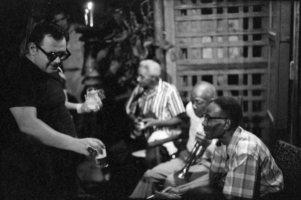 A black-and-white photo showing four people gathered casually. One person stands holding drinks, while three others sit and engage in conversation. They appear relaxed in an indoor setting with dim lighting and a wooden lattice backdrop.