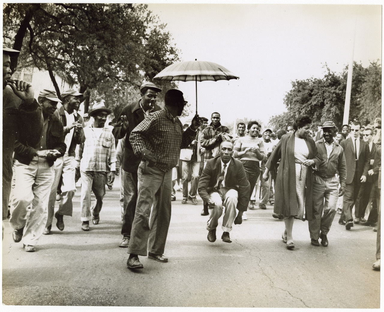 A historic black-and-white photograph features a diverse group of people marching down a tree-lined street. Some individuals hold hands, others carry an umbrella. They appear determined, conveying a sense of unity and purpose.