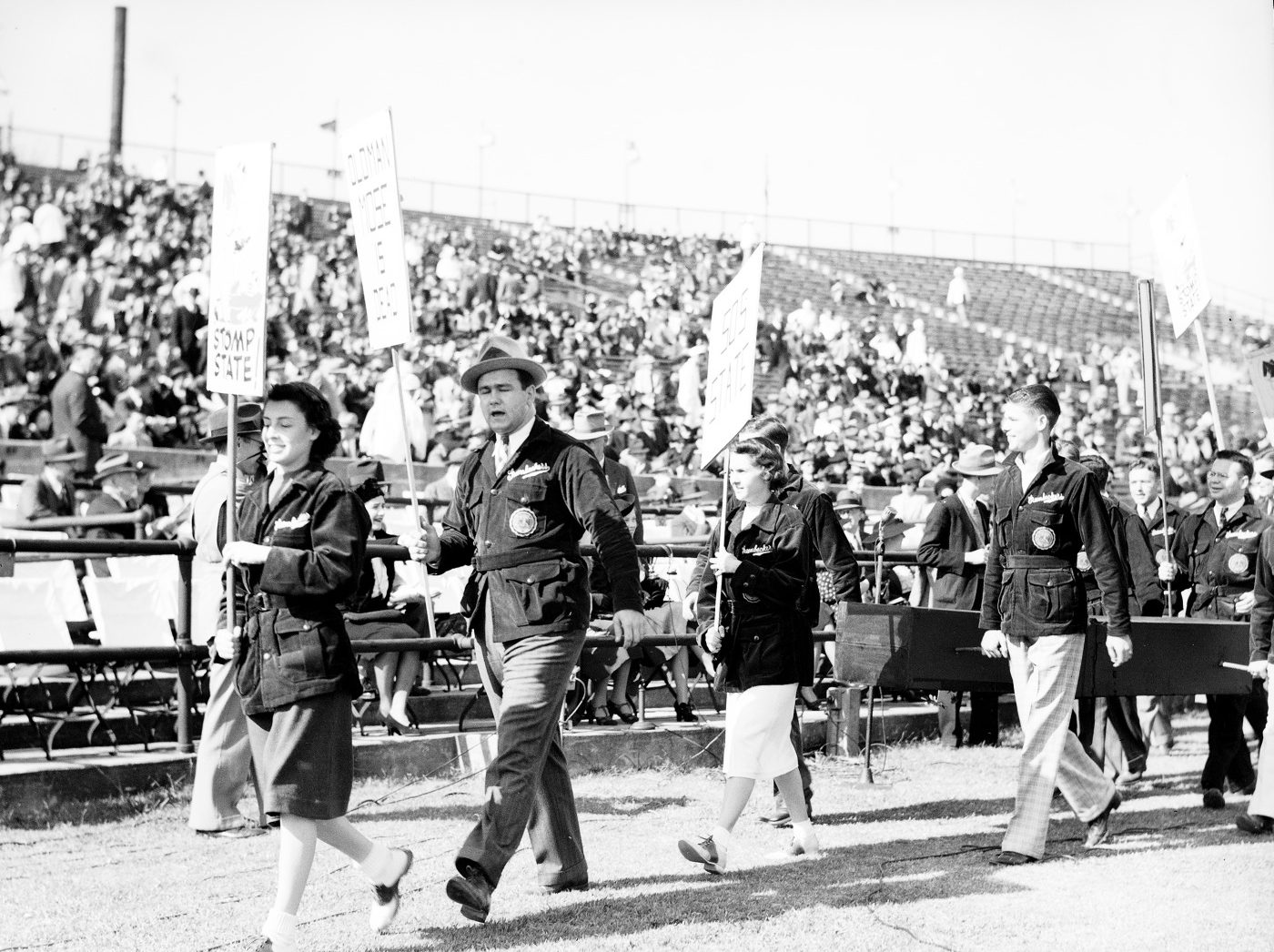 Photo of young women and men carrying signs and wearing jackets. They walk along the sidelines. Spectators can be seen in the stadium stands.