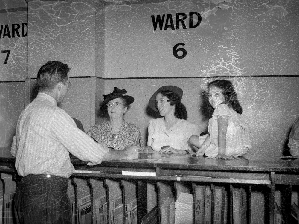 A man in a striped shirt talks to three females at a counter labeled Ward 6. The women, wearing hats, and a young girl, look attentive. The setting suggests a hospital or similar institution. The photo has a vintage, worn appearance.