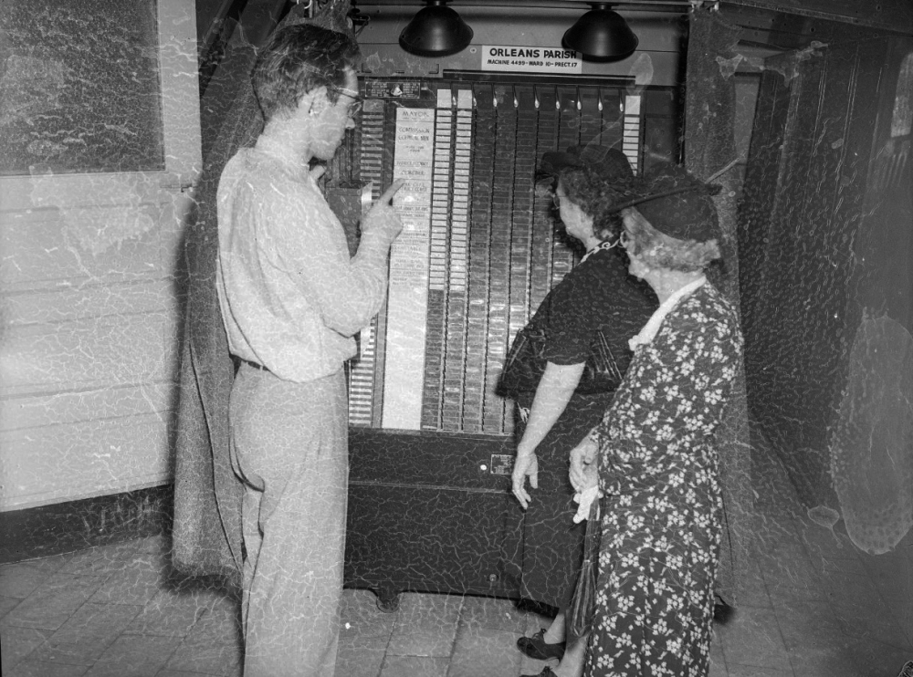 A black-and-white photo shows three people standing in front of a vintage voting machine labeled Orleans Parish. One man points at the machine, while two women dressed in mid-20th-century attire observe. The scene depicts historical voting.