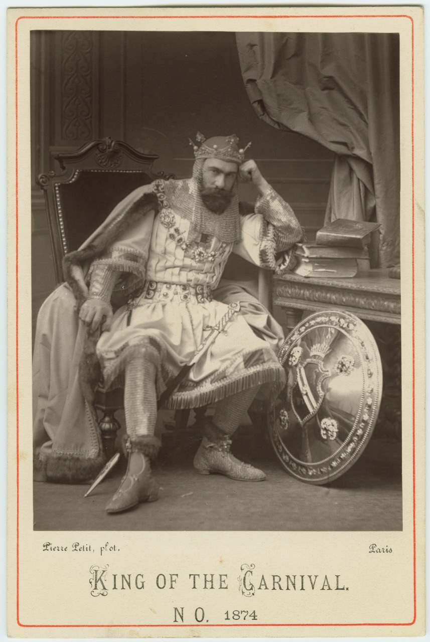 A man dressed as a medieval king sits on a chair, leaning on a table with books and a shield, wearing a crown and ornate robe. The photo is labeled King of the Carnival No. 1874 at the bottom.