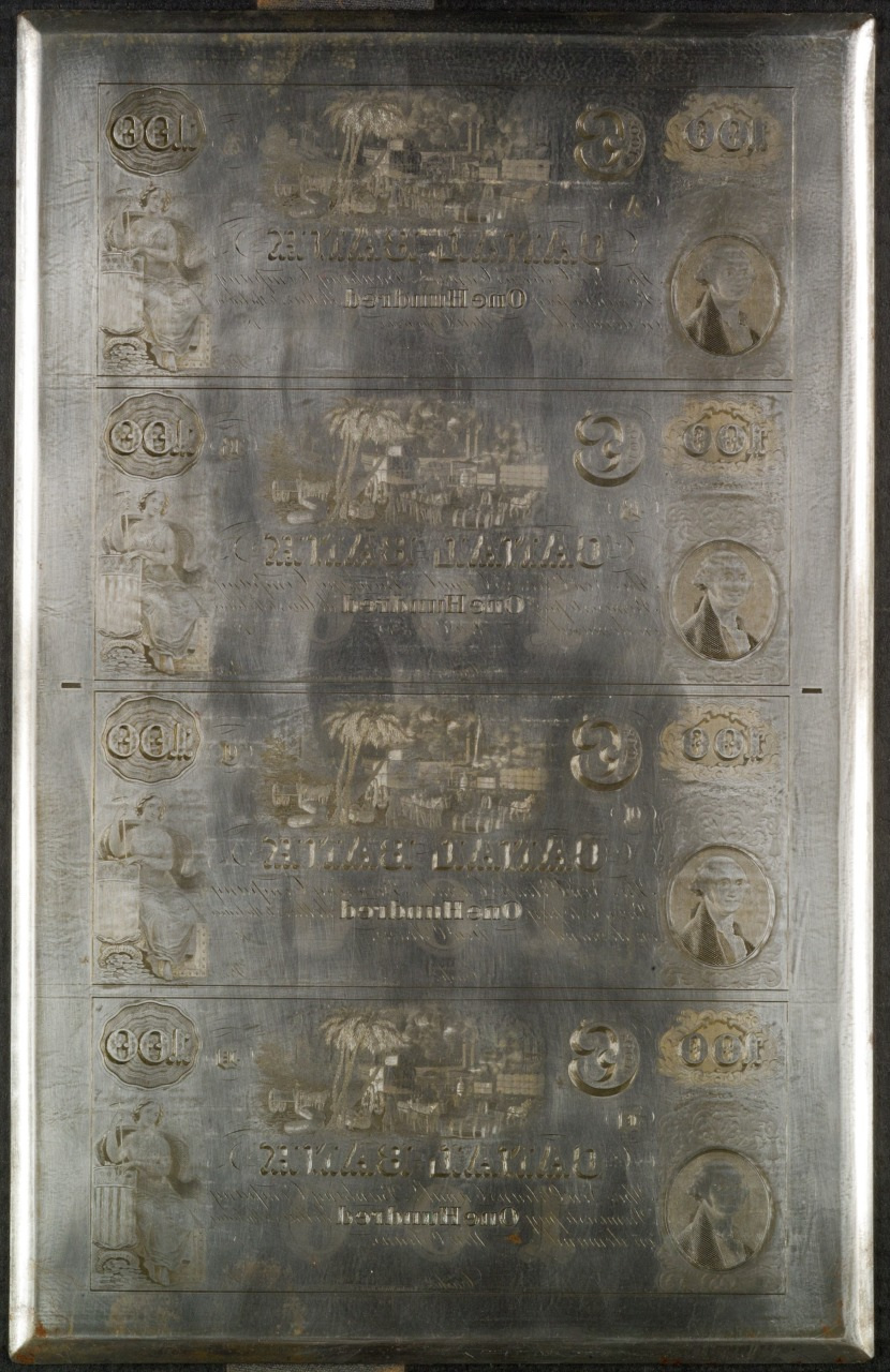 An engraved printing plate featuring four identical designs, possibly for an old currency note. Each section displays intricate illustrations including a seated figure and a circular emblem. The plate is metallic with visible etchings.
