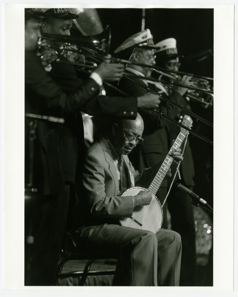 Black and white photo of a seated musician playing a banjo, surrounded by standing brass instrument players in formal attire. The focus is on the banjo player in the foreground, while the brass musicians perform energetically in the background.