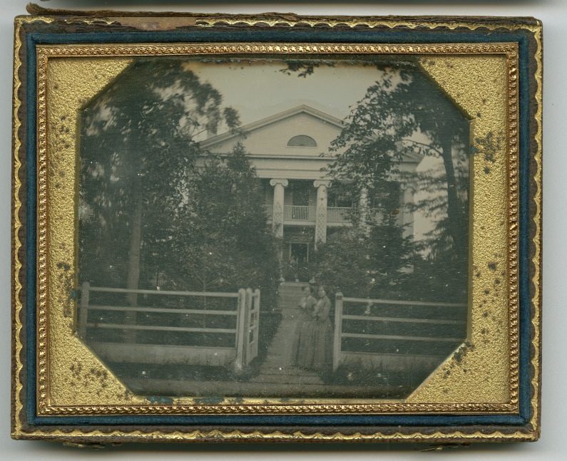 A vintage photograph in an ornate frame shows a large two-story house with columns surrounded by trees. Two figures stand near a wooden fence in the foreground. The image has a sepia tone, indicating its age.