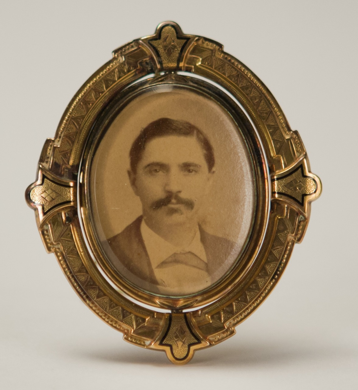 A sepia-toned vintage photograph of a man with a mustache in formal attire, set in an ornate, oval gold frame with decorative engravings, against a neutral background.