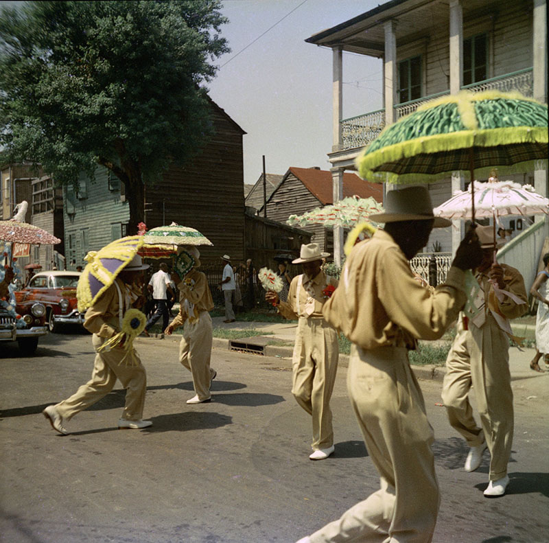 A lively street parade with people in matching tan outfits and hats, carrying colorful parasols. A vintage car is parked in the background, with wooden houses and spectators lining the street.