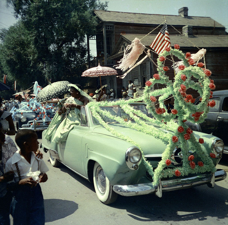 A vintage parade scene with a decorated car adorned with flowers and an American flag. People, including children, watch as the car drives by. One passenger holds a parasol. The setting is a lively street with onlookers and other decorated vehicles.