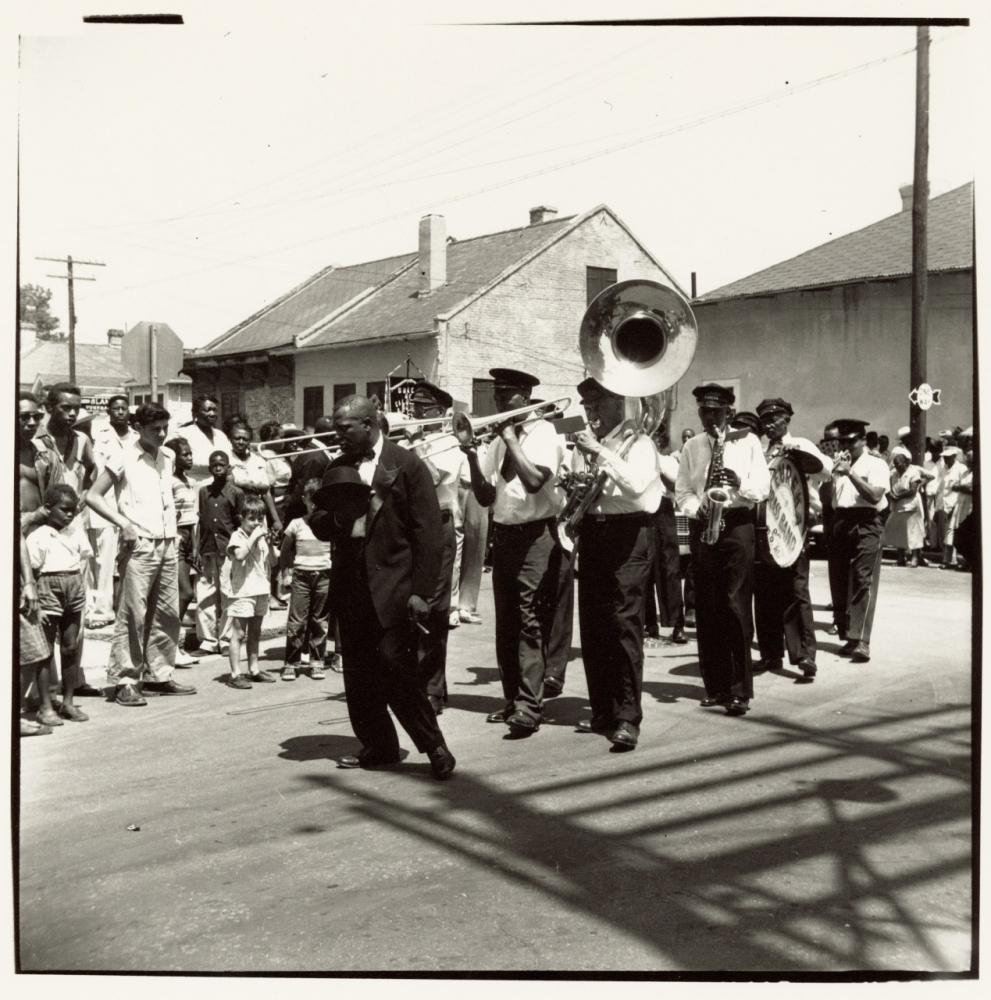 A black and white photo shows a lively street parade with a marching brass band playing instruments, including a sousaphone and trombone. They are followed by an enthusiastic crowd of onlookers, including adults and children, on a sunny day.
