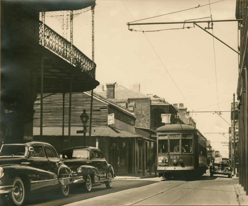 A vintage street scene with a trolley car on tracks and several classic cars parked on the left. The street is lined with old brick buildings and intricate wrought-iron balconies. The photo has a nostalgic, historical atmosphere.