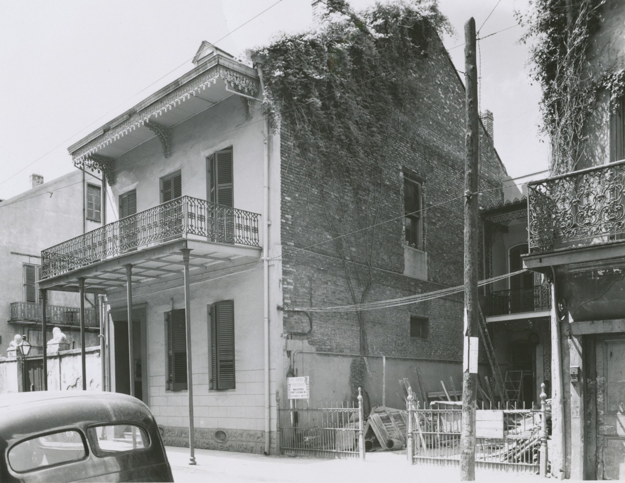 A vintage black-and-white photo shows a two-story building with a balcony and shutters. The side wall is covered in ivy. In front, theres a parked car and a small fenced area with various materials.
