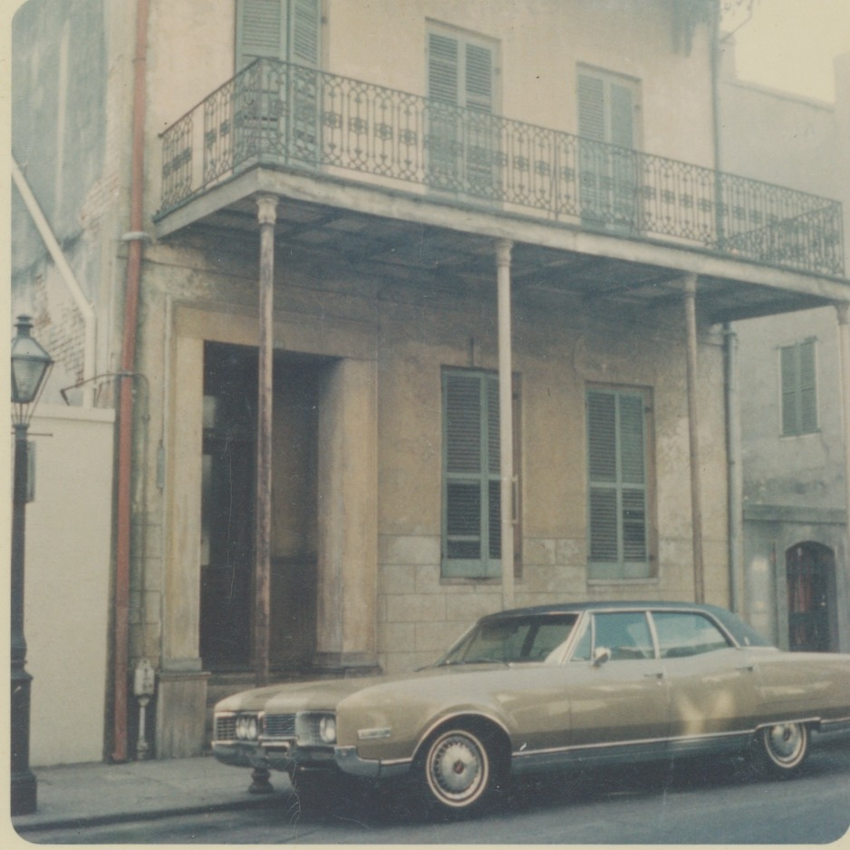 A vintage beige car is parked on a street in front of an old building with a wrought-iron balcony and shuttered windows, showcasing a historic urban scene.