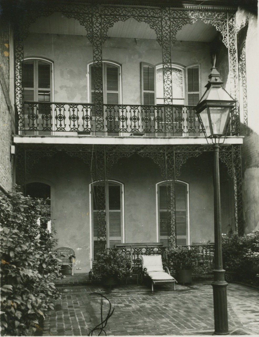 A black and white image of a two-story building with ornate wrought iron balconies. The courtyard features a lounge chair, potted plants, and a vintage street lamp on a brick patio. The shutters on the windows are closed.