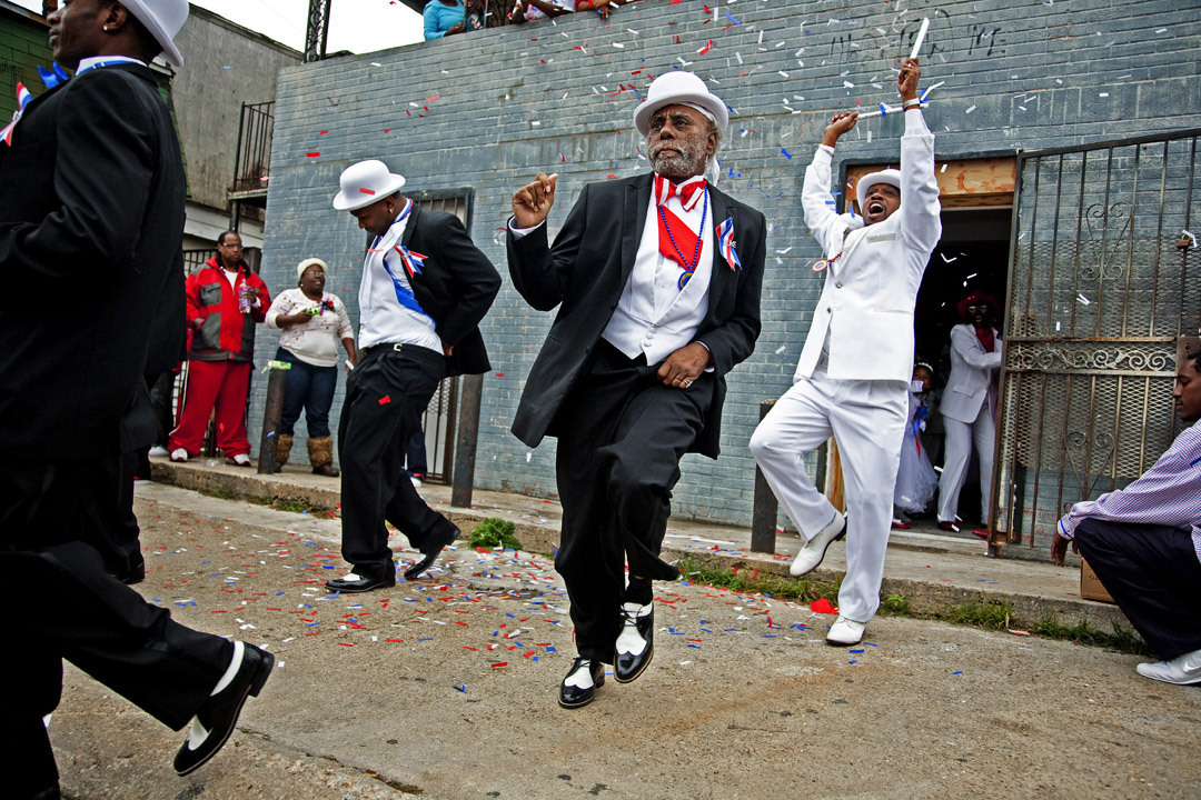 Joyful dancers in formal attire perform energetically on the street, surrounded by colorful confetti. A man in a black suit and hat leads, while another in white joins exuberantly. Onlookers watch from the background.
