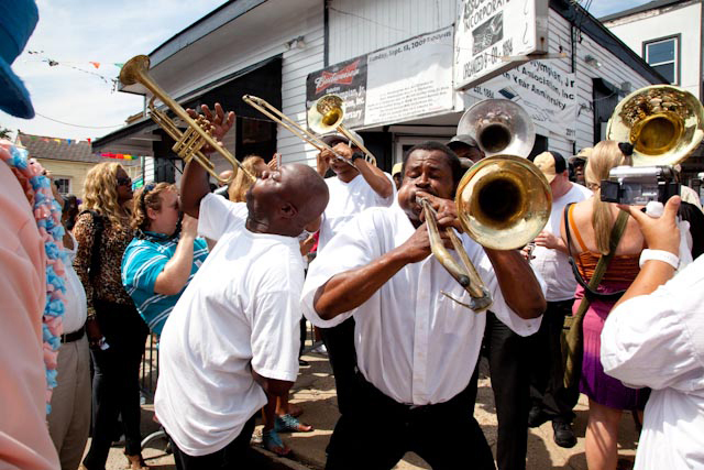 Musicians passionately play brass instruments in a lively outdoor gathering. A man with a trombone and another with a trumpet lead, surrounded by an enthusiastic crowd. The scene is vibrant, with different people enjoying the music.