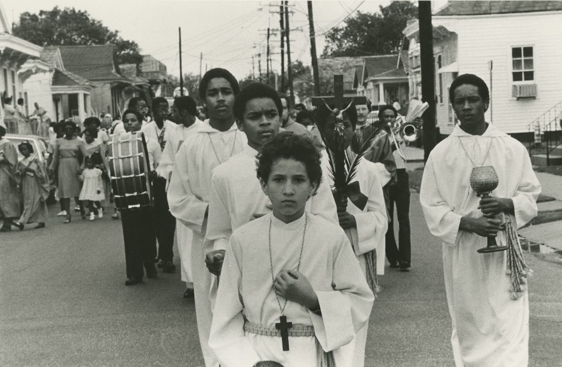 A boy in white lay garments leads a church procession down the street, followed by other boys in white lay garments wearing crucifixes and holding chalices.