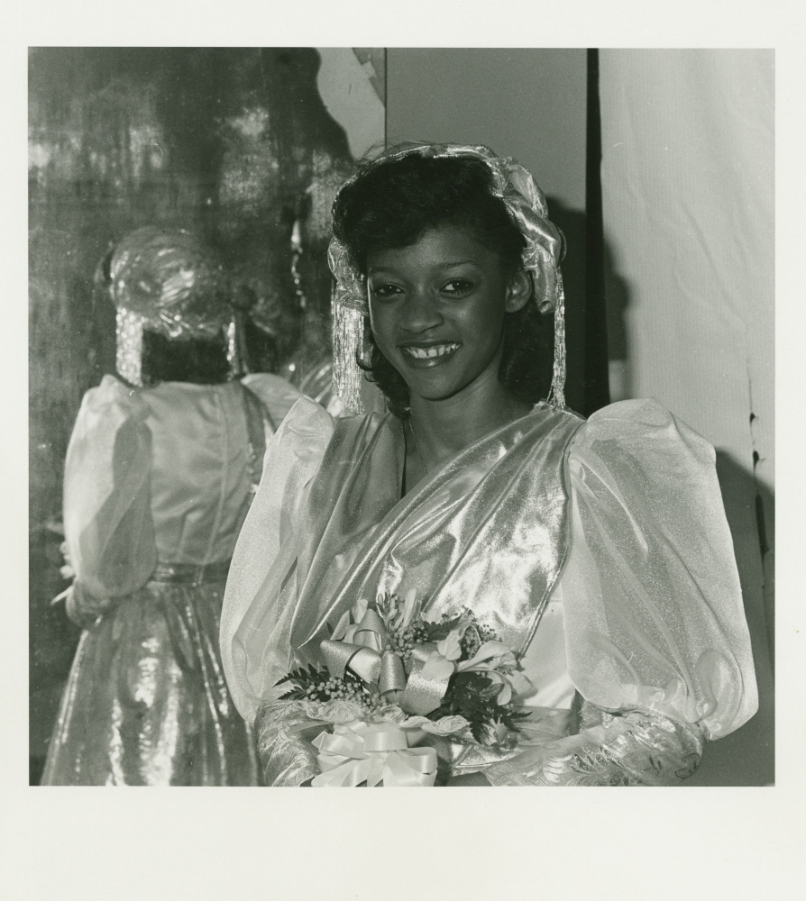 A woman in a shiny dress with puffed sleeves and a veil holds a floral bouquet. She is smiling in front of a reflective surface that shows her back. The setting appears to be indoors.