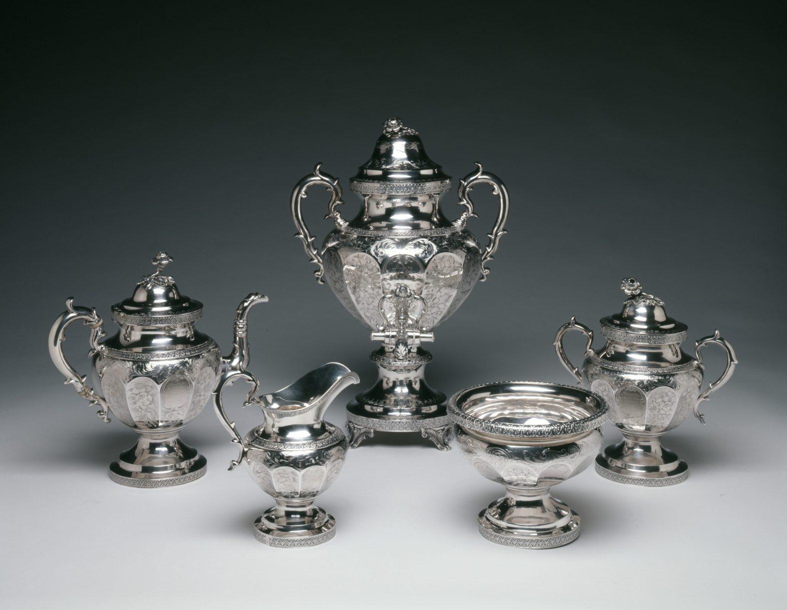 A set of five ornate silver tea service pieces, including a large urn, two teapots, a creamer, and a sugar bowl, all featuring intricate engravings and decorative handles, arranged on a light gray surface against a dark background.