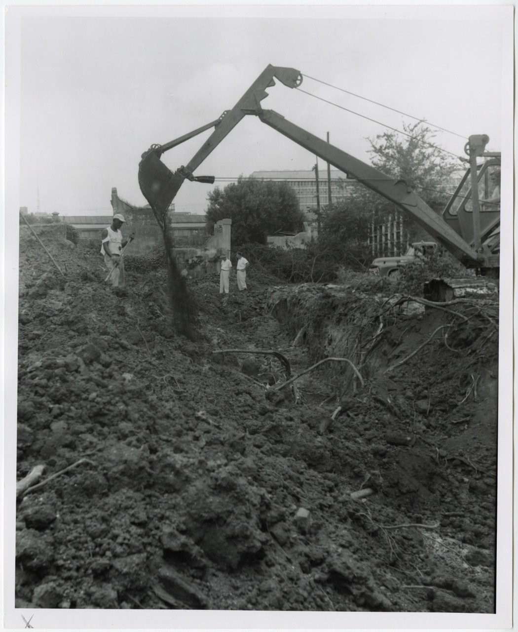An excavator lifts dirt at a construction site, while a group of people in white coats observe nearby. The area is surrounded by trees and distant buildings are visible under a cloudy sky.