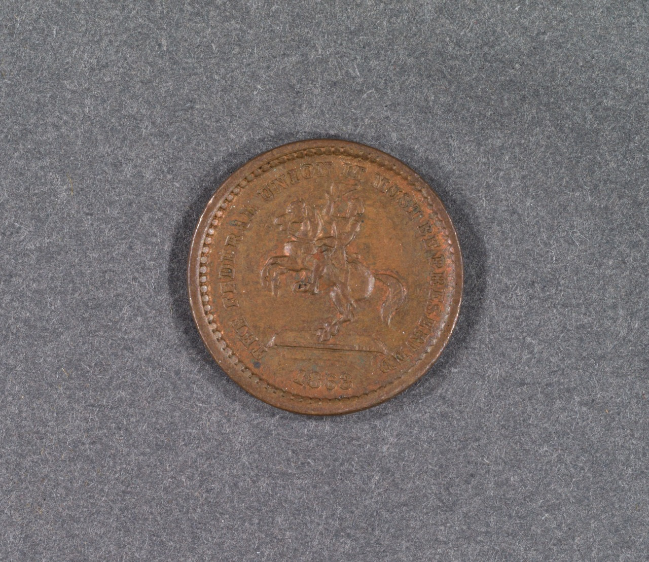 Close-up of a round copper coin featuring a detailed engraving of a knight on horseback. The edges are inscribed with text, and the year 1953 is visible at the bottom. The coin rests on a gray textured surface.