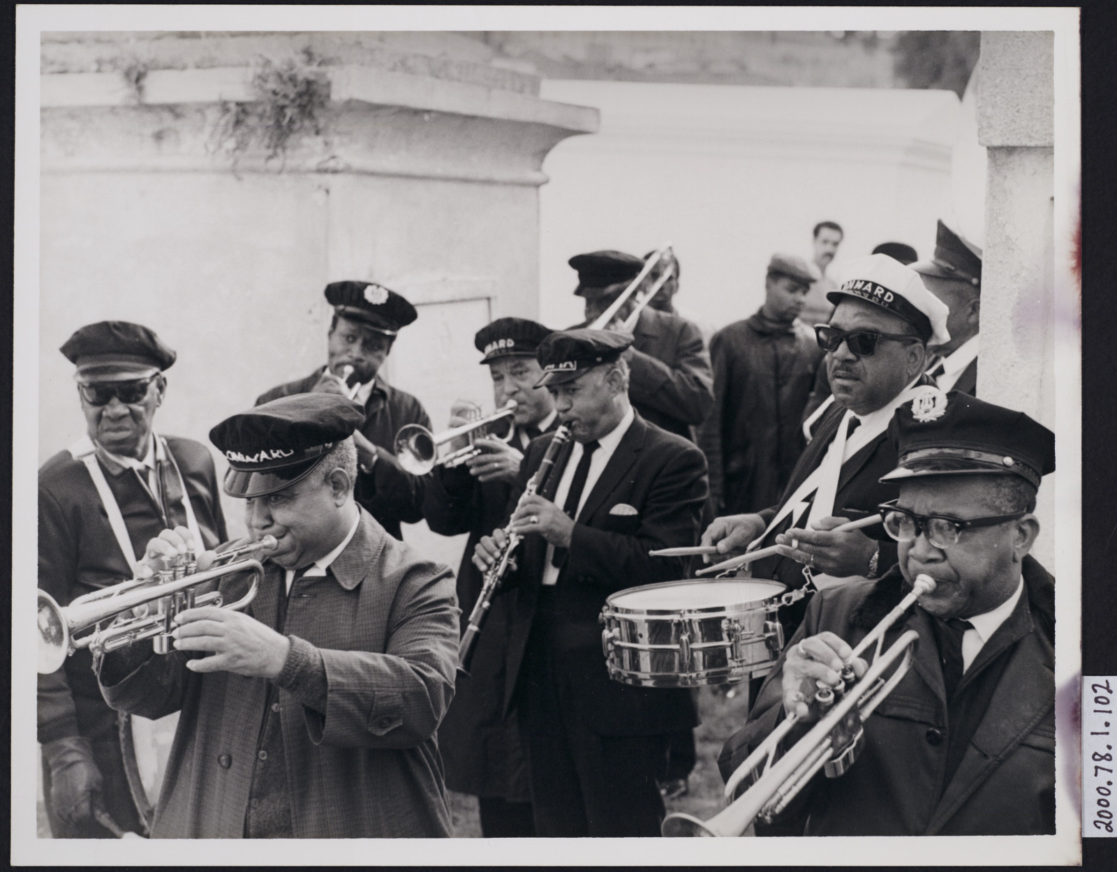 A black-and-white photo of a band performing outdoors. Musicians play trumpets, trombones, and a snare drum, wearing hats and jackets. A small crowd watches in the background. The atmosphere appears lively and engaging.