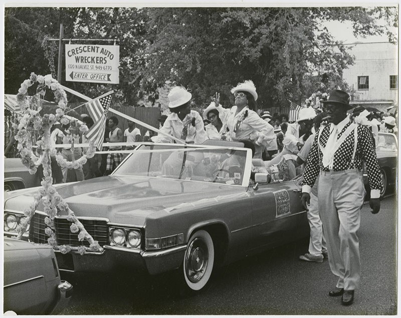 A parade scene featuring people in festive attire, with some standing in a decorated convertible car. An African American man walks beside the car wearing a polka dot shirt. A sign for Crescent Auto Wreckers is visible in the background.