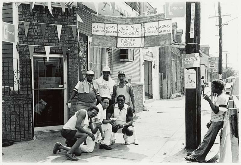 A group of six people pose in front of a brick building with a sign overhead. Some wear hats and athletic attire. A man to the right leans on a pole with posters. The scene is an urban street with a pay phone and weathered buildings.