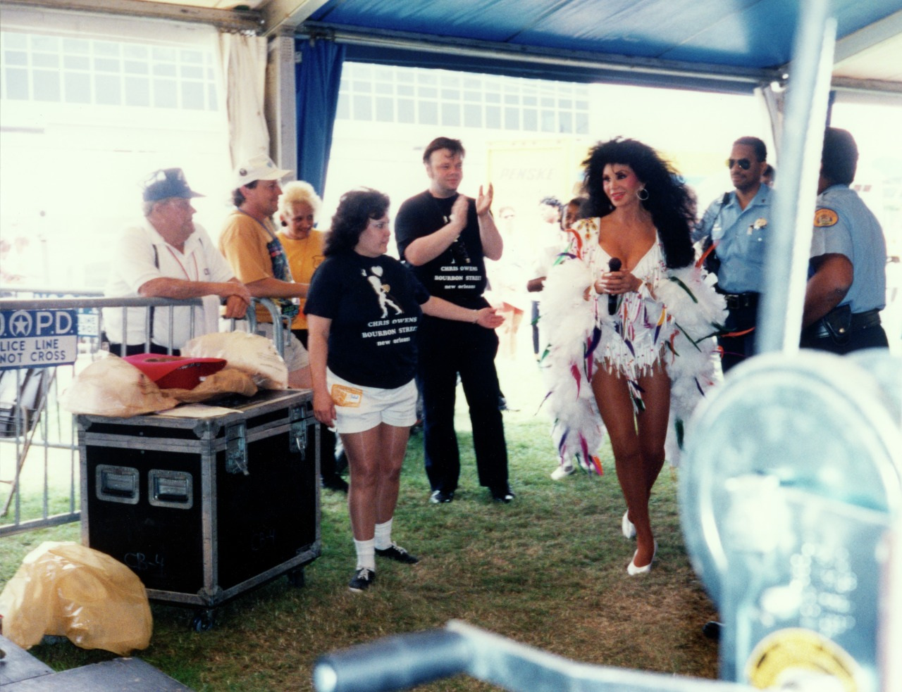 A person in a vibrant feathered costume walks confidently near a crowd under a blue and white tent. Onlookers, including a woman in a black shirt and several security personnel, observe. The scene suggests a lively, festive event.