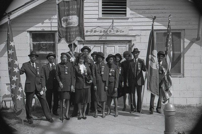 A vintage black and white photo of a group of men and women in formal attire, standing in front of a building. They hold banners and flags. The building has a sign above the door with text. The group seems to be part of an organized event or gathering.