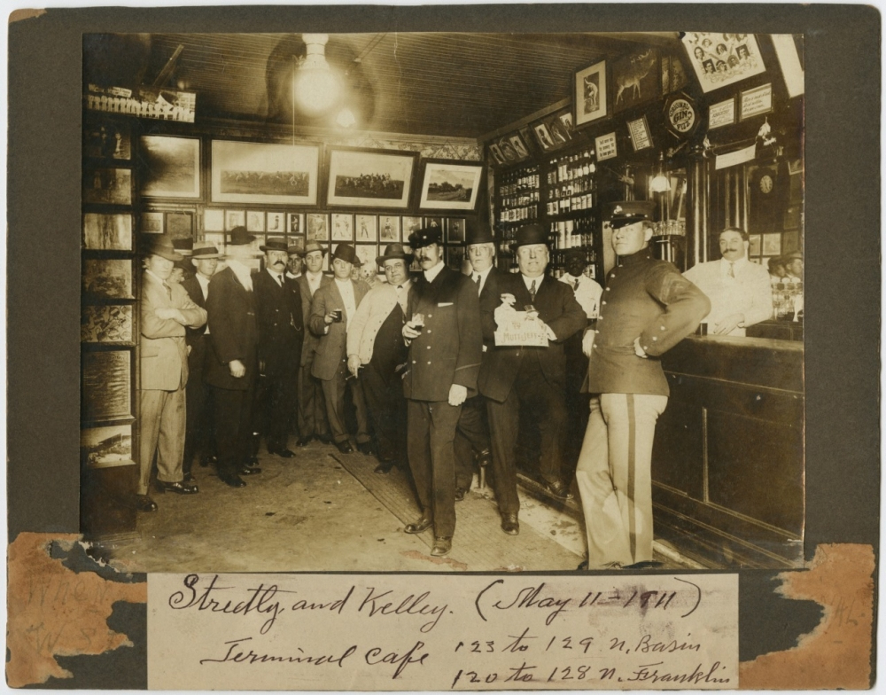 A vintage photo of men in suits and hats gathered in a bar. The bar is lined with framed photos and shelves of bottles. A bartender stands behind the counter. Handwritten text below reads Streets and Kelley, Terminal Café, May 11, 1911, 123 to 127 N. Basin.